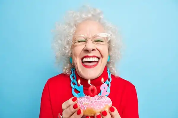 Happy birthday overjoyed senior lady smiles broadly has white perfect teeth going to blow candles on glazed doughnuts being well dressed celebrates 102nd bday enjoys party with relatives - Perspon Centred Care Training Course