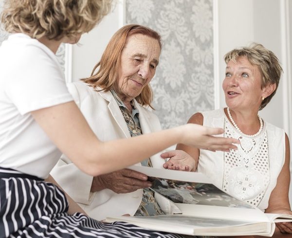 People gathered around a photograph album, smiling and gesturing towards each other. Life Story Work training. Cognitive Stimulation Therapy.