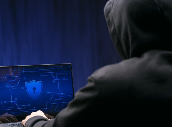 Hooded person sitting in front of a laptop. Security sign on screen. Hacking. Prevent training.